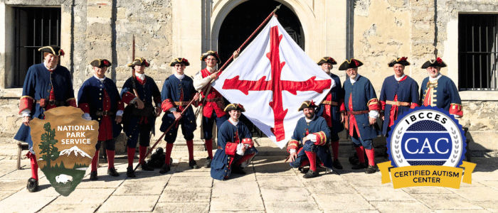 A group of reenactors dressed in historic military uniforms pose with a large cross-emblazoned flag in front of a stone building, with the National Park Service and Certified Autism Center™ (CAC) logos displayed on either side.