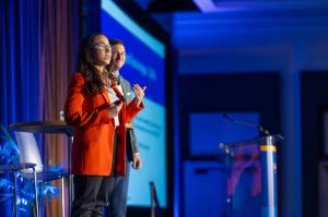 Woman in an orange jacket addresses conference attendees from the main stage